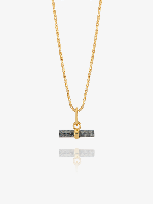 A gold chain necklace with a rectangular dark gray stone pendant featuring a gold split ring and clasp, displayed against a light gray background with a soft reflection below.