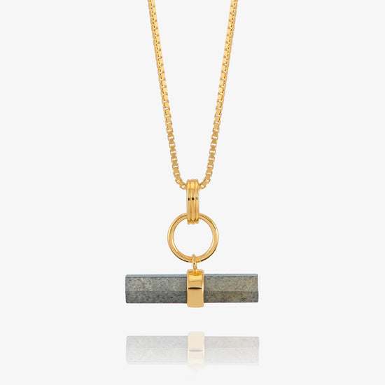 A gold chain necklace with a horizontal bar pendant featuring a dark gray stone inlay and a gold accent, shown against a light gray background with a soft reflection below.