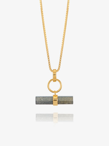 A gold chain necklace with a horizontal bar pendant featuring a dark gray stone inlay and a gold accent, shown against a light gray background with a soft reflection below.