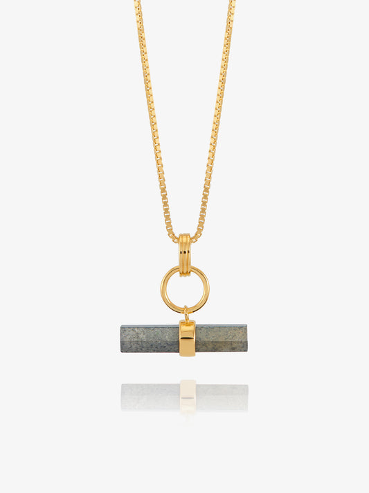 A gold chain necklace with a horizontal bar pendant featuring a dark gray stone inlay and a gold accent, shown against a light gray background with a soft reflection below.