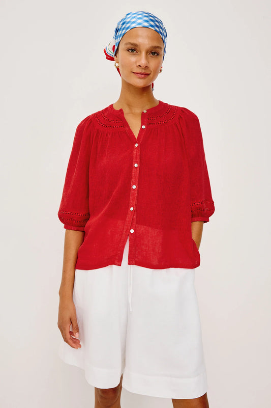 Woman wearing a red blouse and white shorts with a blue and white checkered headscarf on a light background