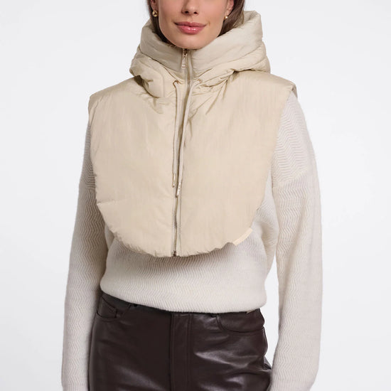 A woman modeling a beige hooded puffer vest with a wide collar over a cream knit sweater and dark brown pants, shot against a plain light gray studio background.
