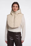 A woman modeling a beige hooded puffer vest with a wide collar over a cream knit sweater and dark brown pants, shot against a plain light gray studio background.