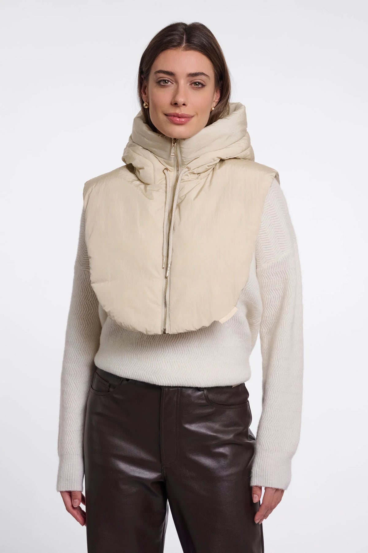 A woman modeling a beige hooded puffer vest with a wide collar over a cream knit sweater and dark brown pants, shot against a plain light gray studio background.