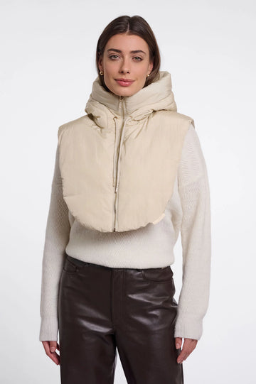 A woman modeling a beige hooded puffer vest with a wide collar over a cream knit sweater and dark brown pants, shot against a plain light gray studio background.
