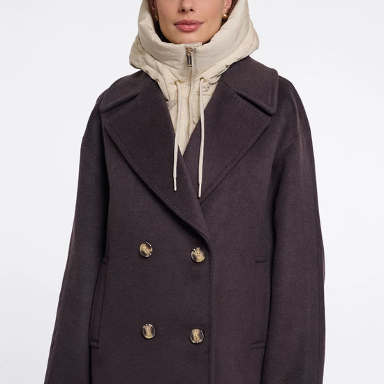 A woman wearing a dark charcoal double-breasted coat over a cream zip-up hoodie, photographed against a plain light gray studio background.
