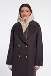 A woman wearing a dark charcoal double-breasted coat over a cream zip-up hoodie, photographed against a plain light gray studio background.