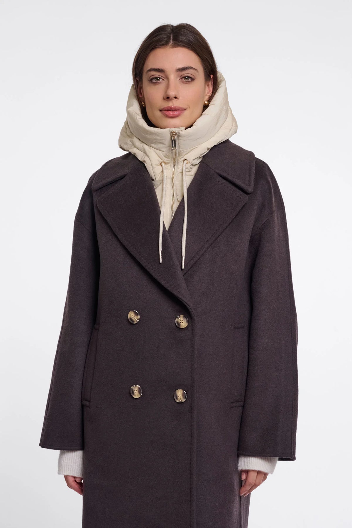 A woman wearing a dark charcoal double-breasted coat over a cream zip-up hoodie, photographed against a plain light gray studio background.