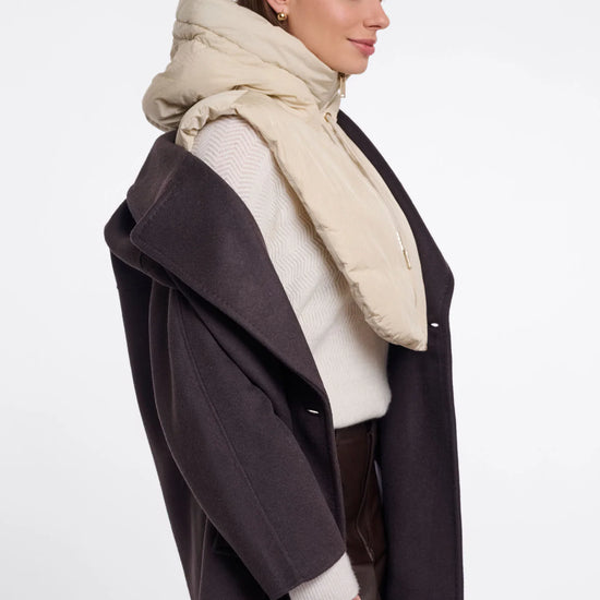 A woman in profile wearing a dark brown coat, cream knit sweater, and a thick beige puffer scarf against a plain light gray studio background.