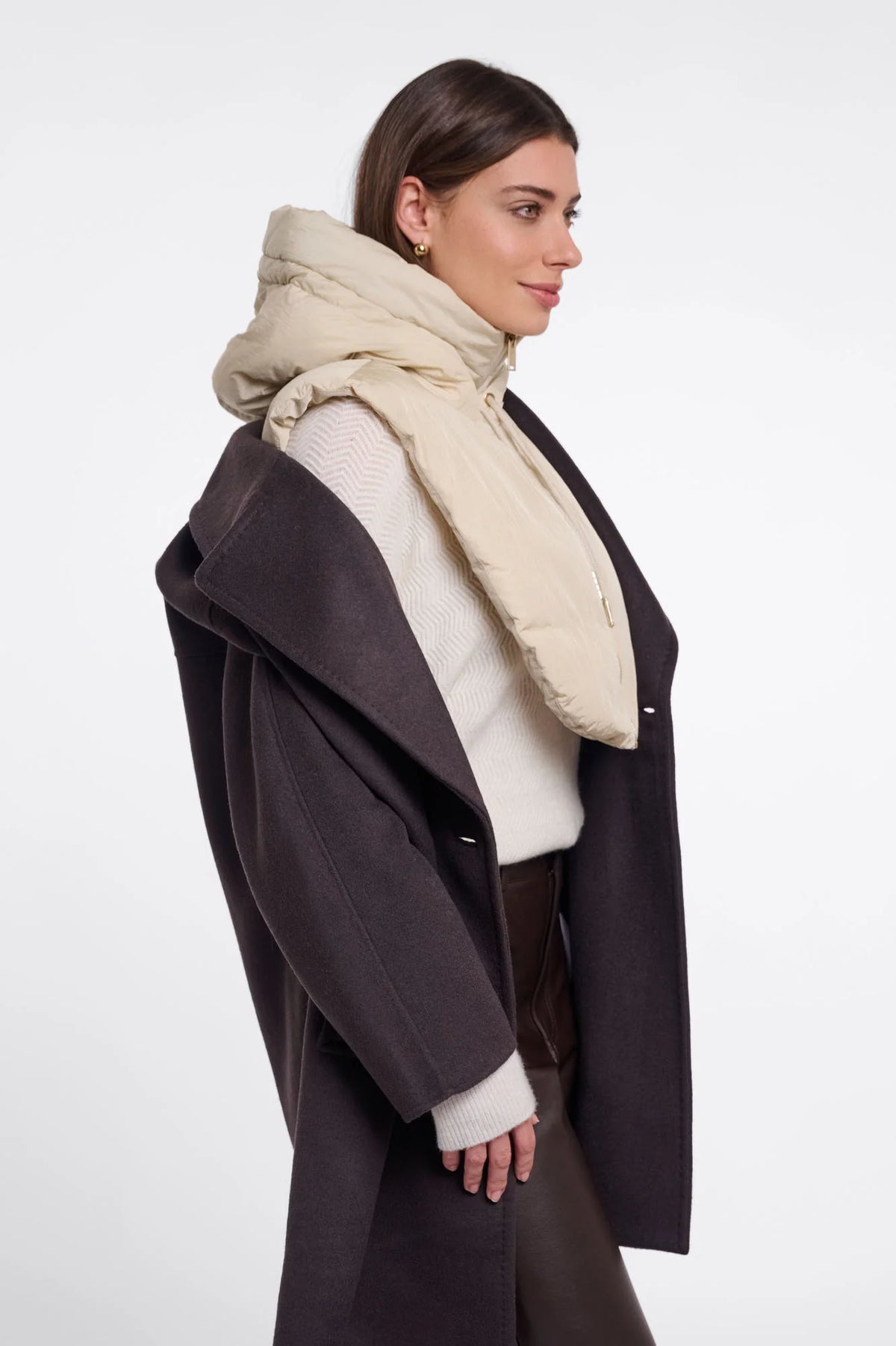 A woman in profile wearing a dark brown coat, cream knit sweater, and a thick beige puffer scarf against a plain light gray studio background.