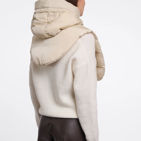 A woman shown from the back wearing a cream-colored knit sweater with a large, padded quilted hood attached, paired with dark brown pants, against a plain white studio background.