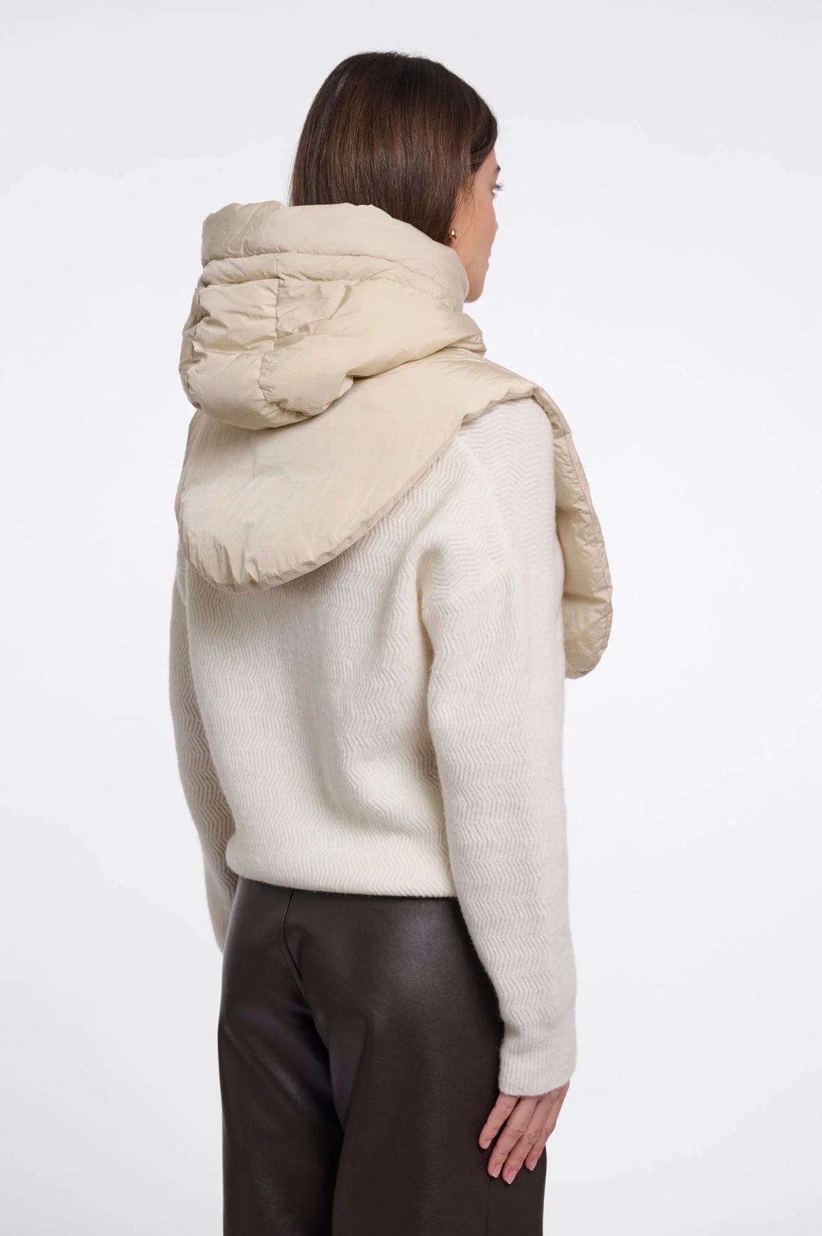A woman shown from the back wearing a cream-colored knit sweater with a large, padded quilted hood attached, paired with dark brown pants, against a plain white studio background.