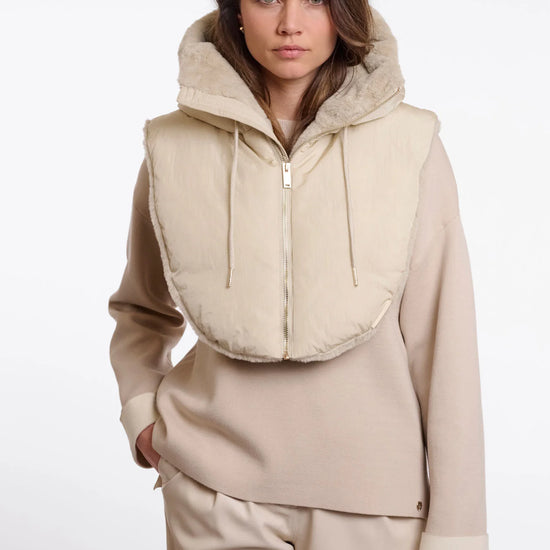 A model wearing a beige hooded zip-up vest with a soft fuzzy trim over a matching beige long-sleeve top and trousers, shot against a plain light gray studio background.
