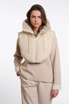 A model wearing a beige hooded zip-up vest with a soft fuzzy trim over a matching beige long-sleeve top and trousers, shot against a plain light gray studio background.