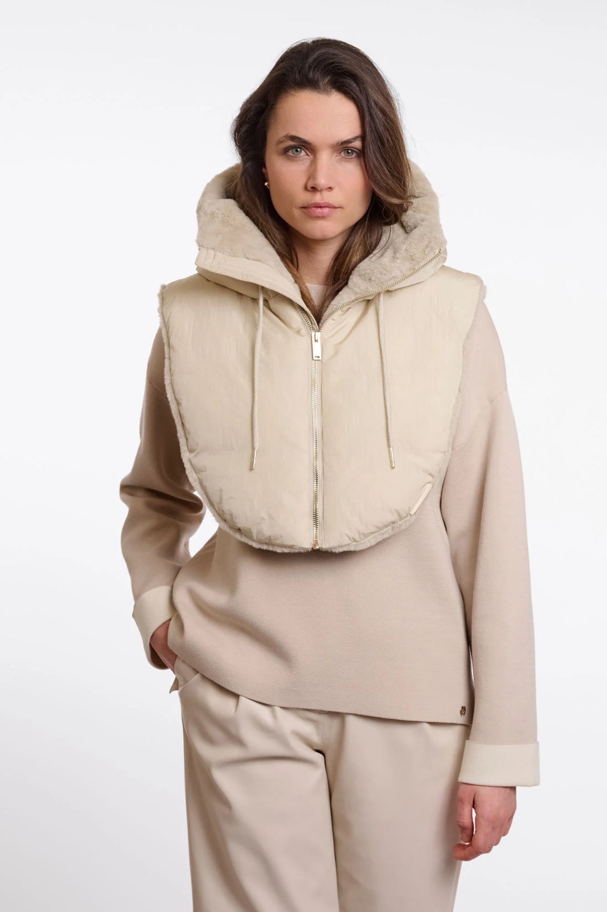 A model wearing a beige hooded zip-up vest with a soft fuzzy trim over a matching beige long-sleeve top and trousers, shot against a plain light gray studio background.