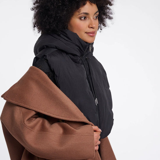 A woman with curly black hair is shown in profile wearing a black puffer jacket and holding a brown coat against a plain light gray studio background.