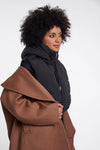 A woman with curly black hair is shown in profile wearing a black puffer jacket and holding a brown coat against a plain light gray studio background.