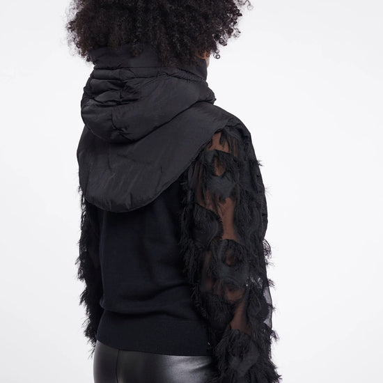A model shown from the back wearing a black puffer jacket with textured, feather-like sleeves over a black top and shiny black pants, set against a plain white studio background.
