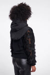 A model shown from the back wearing a black puffer jacket with textured, feather-like sleeves over a black top and shiny black pants, set against a plain white studio background.