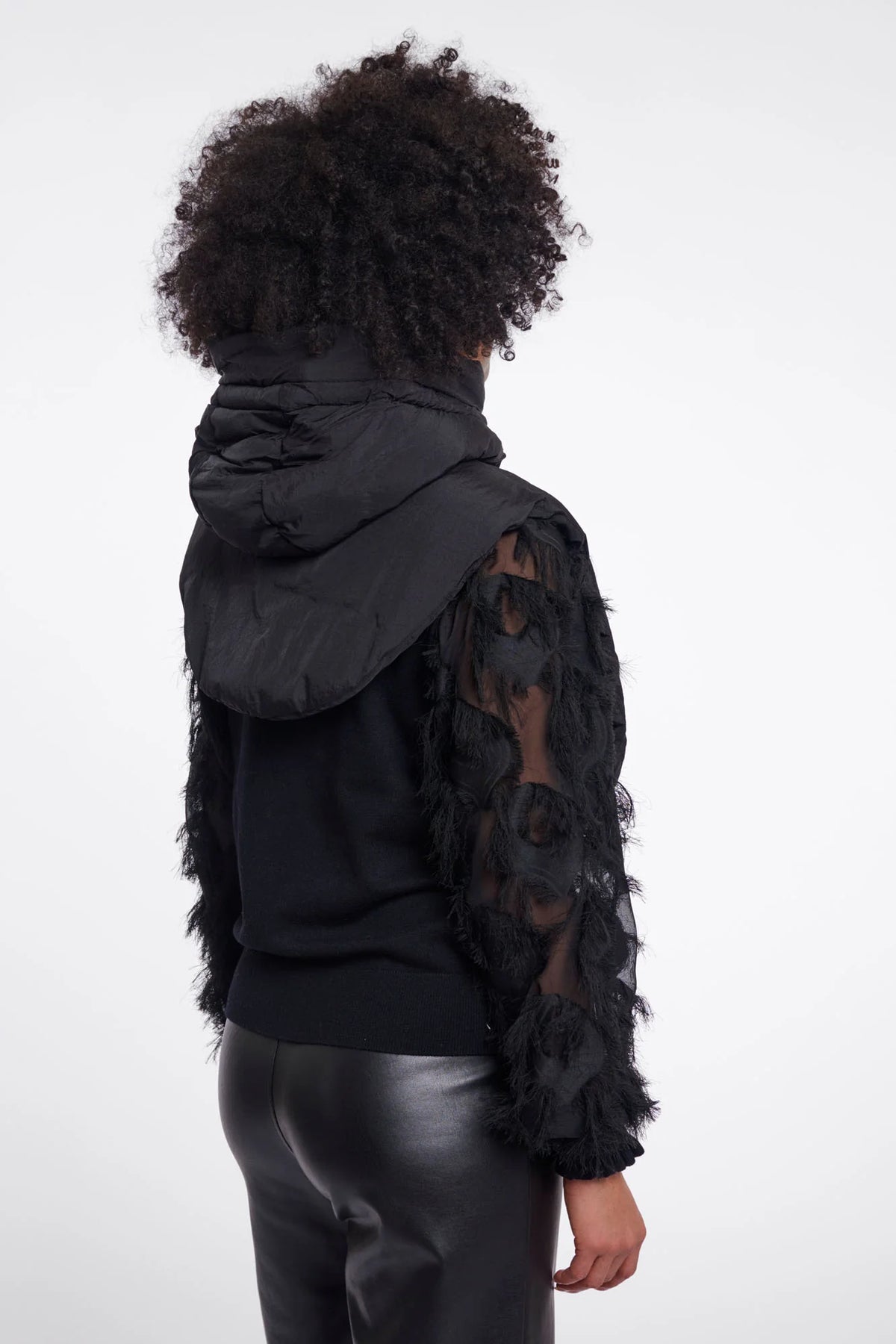 A model shown from the back wearing a black puffer jacket with textured, feather-like sleeves over a black top and shiny black pants, set against a plain white studio background.