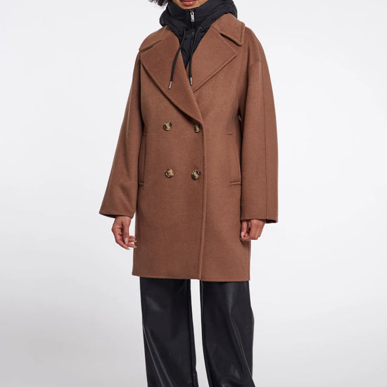 A model wearing a camel brown double-breasted coat over a black puffer jacket and black wide-leg trousers, standing against a light gray studio background.