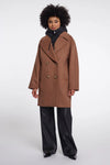 A model wearing a camel brown double-breasted coat over a black puffer jacket and black wide-leg trousers, standing against a light gray studio background.