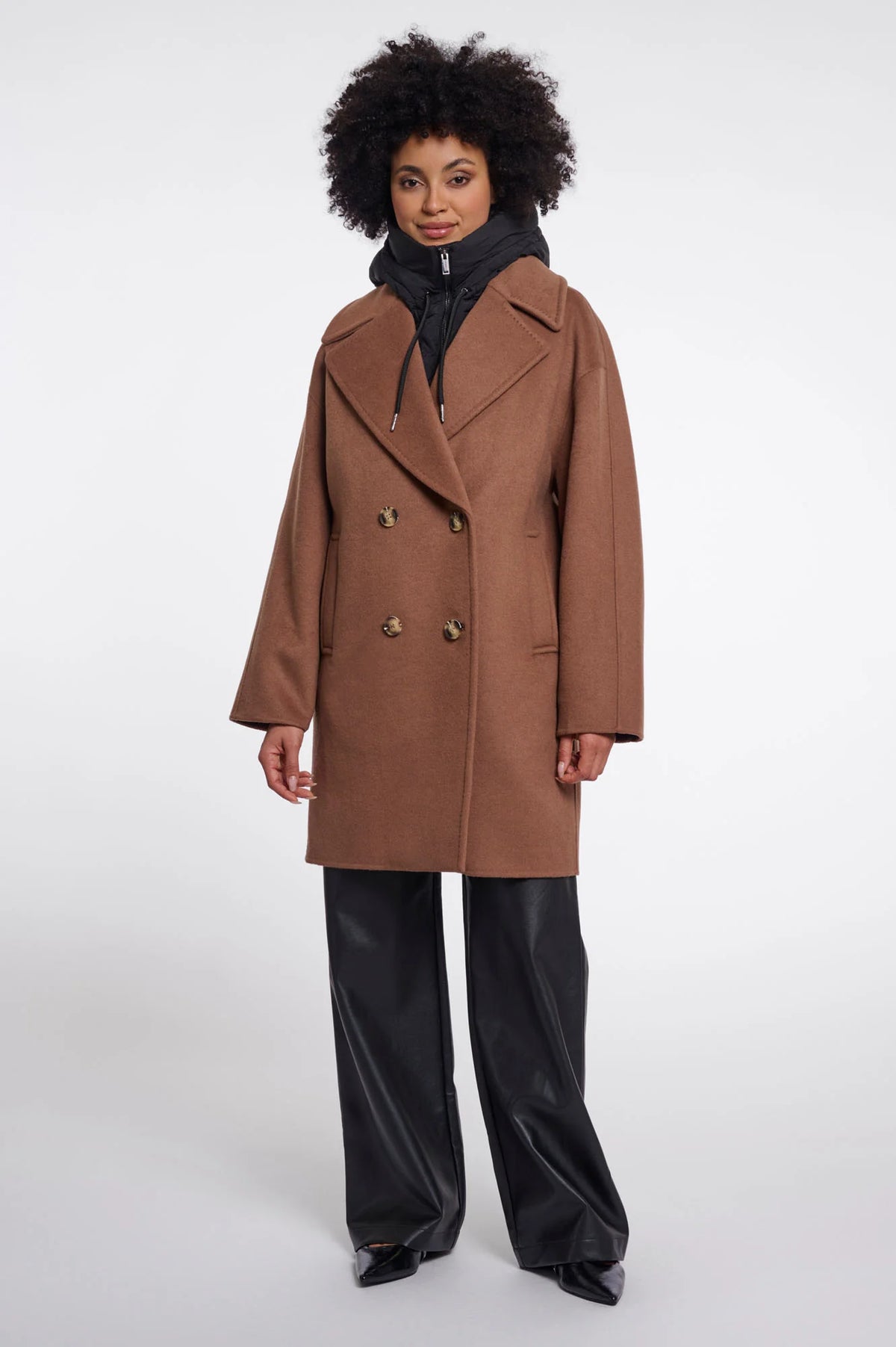 A model wearing a camel brown double-breasted coat over a black puffer jacket and black wide-leg trousers, standing against a light gray studio background.