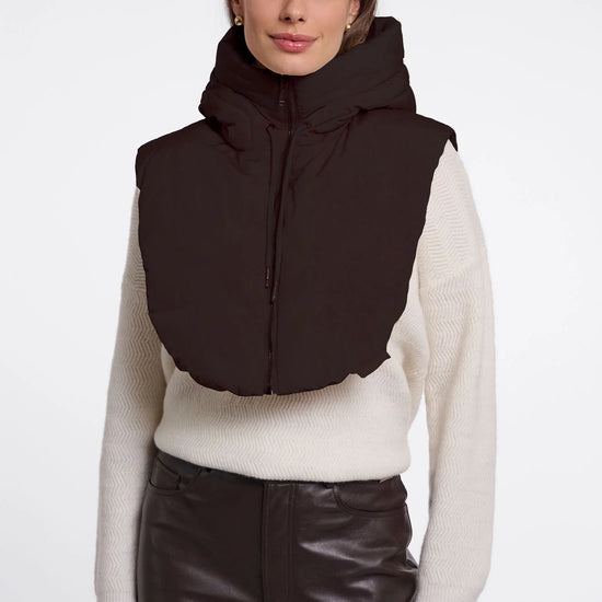 A woman modeling a dark brown puffer vest with a high collar over a cream ribbed sweater and dark brown pants, shot against a plain light gray studio background.