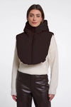 A woman modeling a dark brown puffer vest with a high collar over a cream ribbed sweater and dark brown pants, shot against a plain light gray studio background.