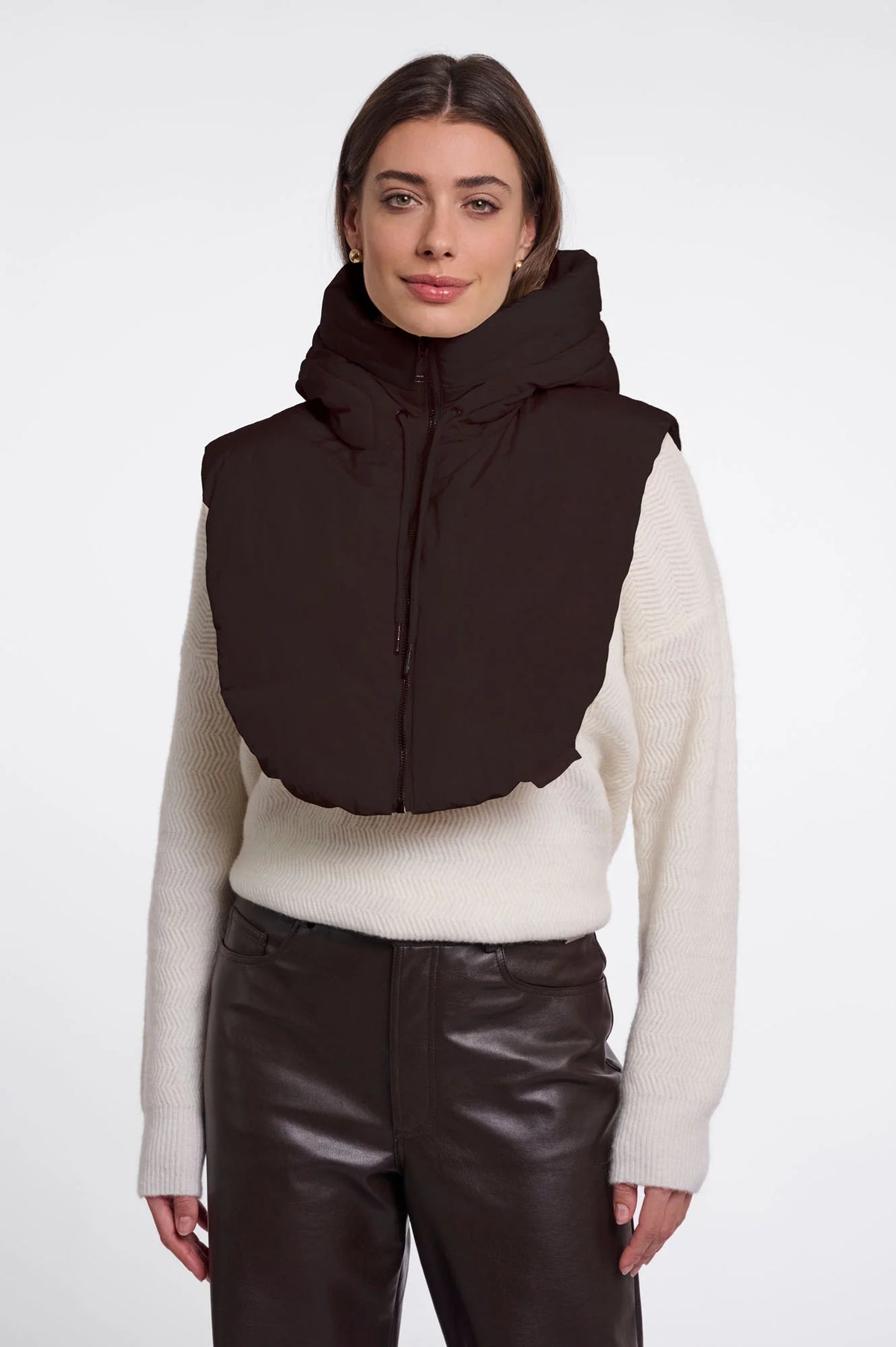 A woman modeling a dark brown puffer vest with a high collar over a cream ribbed sweater and dark brown pants, shot against a plain light gray studio background.