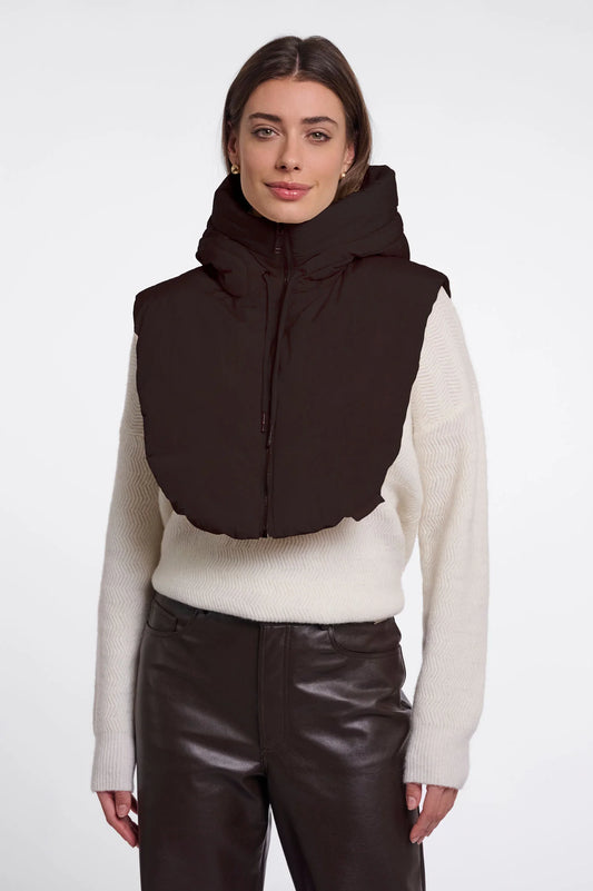 A woman modeling a dark brown puffer vest with a high collar over a cream ribbed sweater and dark brown pants, shot against a plain light gray studio background.