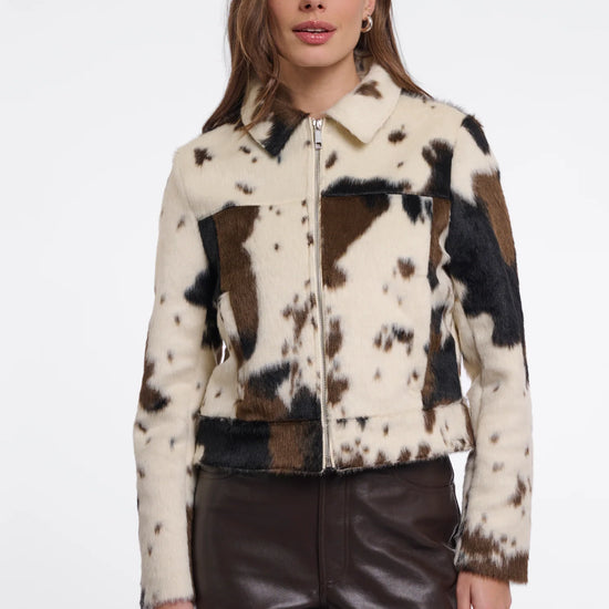 A woman modeling a cropped faux-fur jacket with a cow-print pattern in cream, brown, and black, paired with dark brown faux-leather pants, against a plain white studio background.