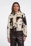A woman modeling a cropped faux-fur jacket with a cow-print pattern in cream, brown, and black, paired with dark brown faux-leather pants, against a plain white studio background.