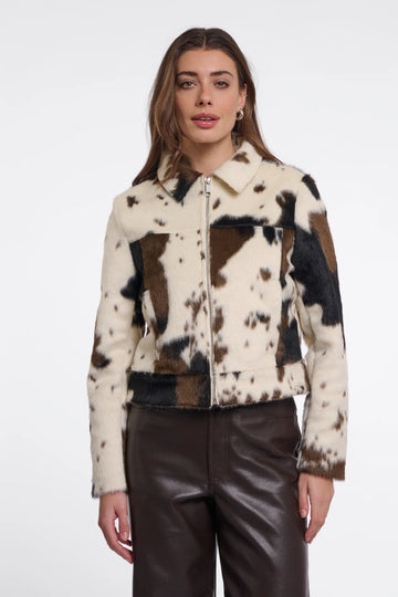 A woman modeling a cropped faux-fur jacket with a cow-print pattern in cream, brown, and black, paired with dark brown faux-leather pants, against a plain white studio background.