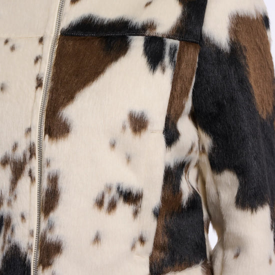 Close-up of a cropped faux-fur jacket with a cow-print pattern in cream, brown, and black, featuring a front zipper and collar.