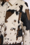 Close-up of a cropped faux-fur jacket with a cow-print pattern in cream, brown, and black, featuring a front zipper and collar.