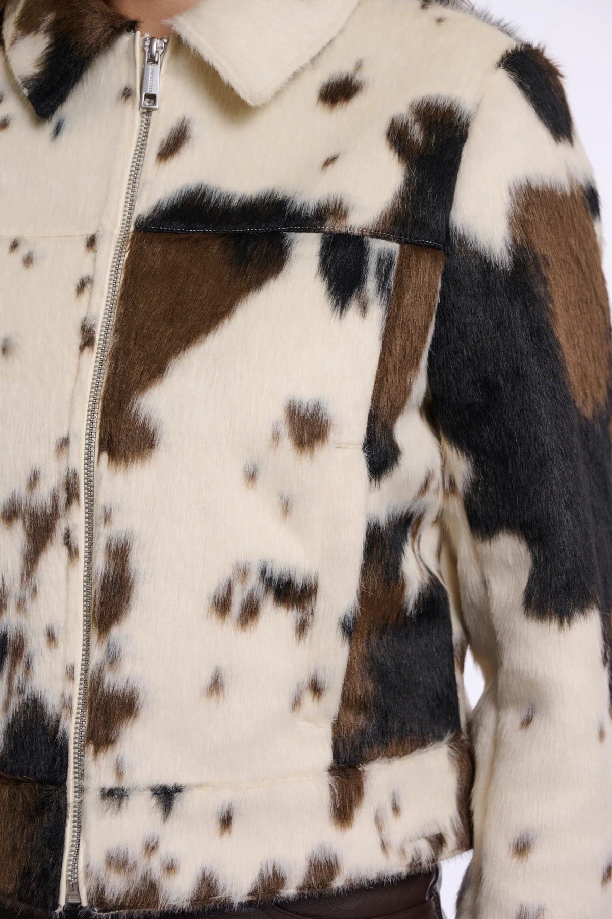 Close-up of a cropped faux-fur jacket with a cow-print pattern in cream, brown, and black, featuring a front zipper and collar.
