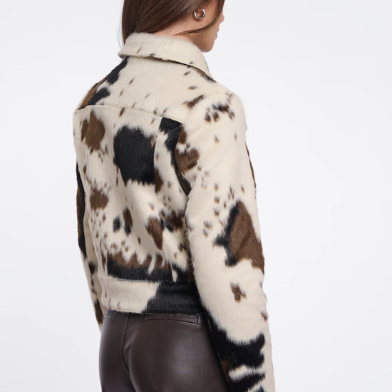 A woman shown from the back wearing a cropped faux-fur jacket with a cow-print pattern in cream, brown, and black, paired with dark brown pants, against a plain white studio background.