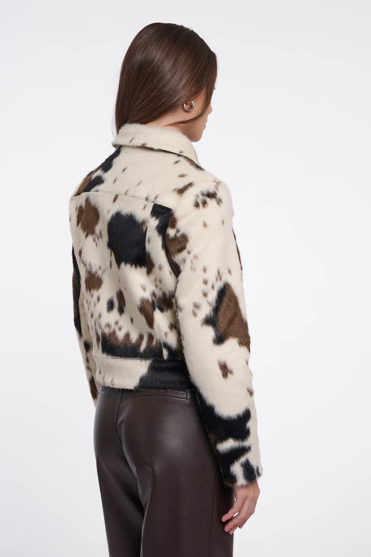 A woman shown from the back wearing a cropped faux-fur jacket with a cow-print pattern in cream, brown, and black, paired with dark brown pants, against a plain white studio background.