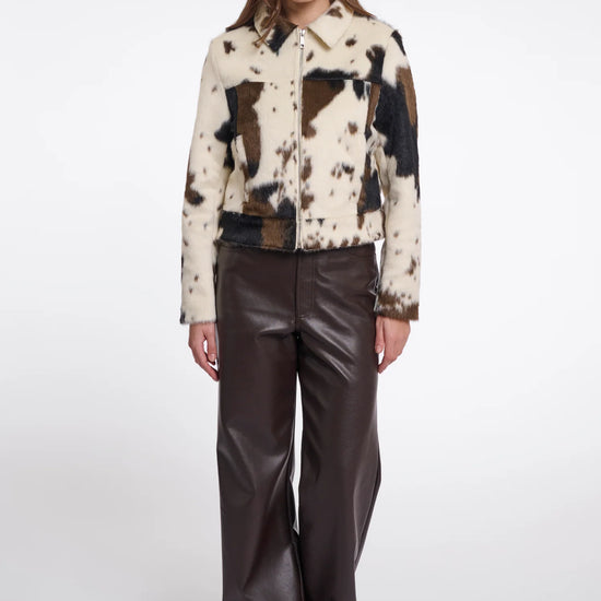 A model stands against a light gray studio background wearing a cropped patchwork jacket with cowhide-style panels in cream, brown, and black, paired with dark brown wide-leg faux-leather pants.