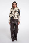 A model stands against a light gray studio background wearing a cropped patchwork jacket with cowhide-style panels in cream, brown, and black, paired with dark brown wide-leg faux-leather pants.