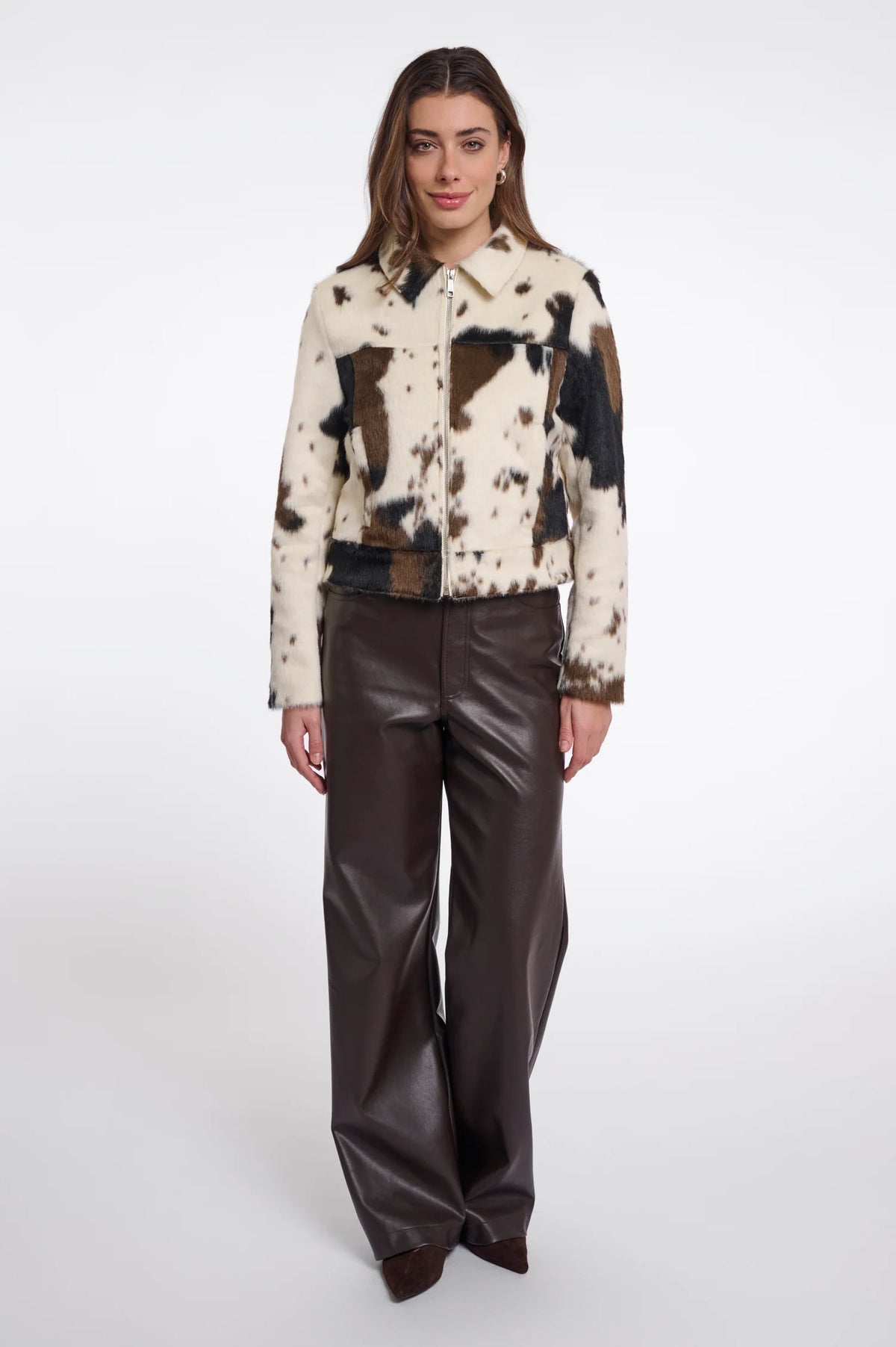 A model stands against a light gray studio background wearing a cropped patchwork jacket with cowhide-style panels in cream, brown, and black, paired with dark brown wide-leg faux-leather pants.