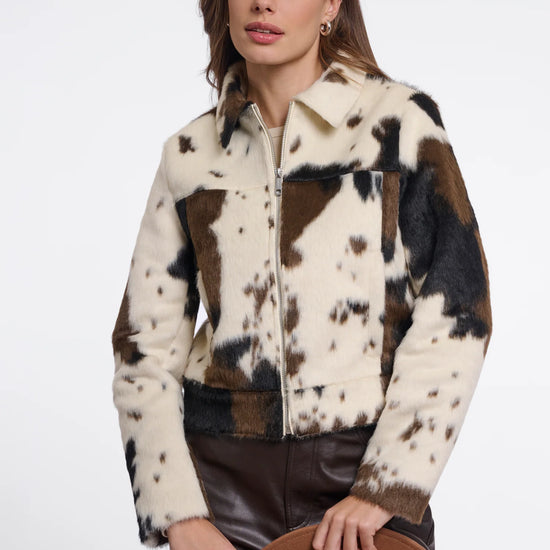 A woman models a cow-print faux fur jacket with brown leather pants and holds a brown wide-brim hat against a plain white studio background.