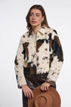 A woman models a cow-print faux fur jacket with brown leather pants and holds a brown wide-brim hat against a plain white studio background.