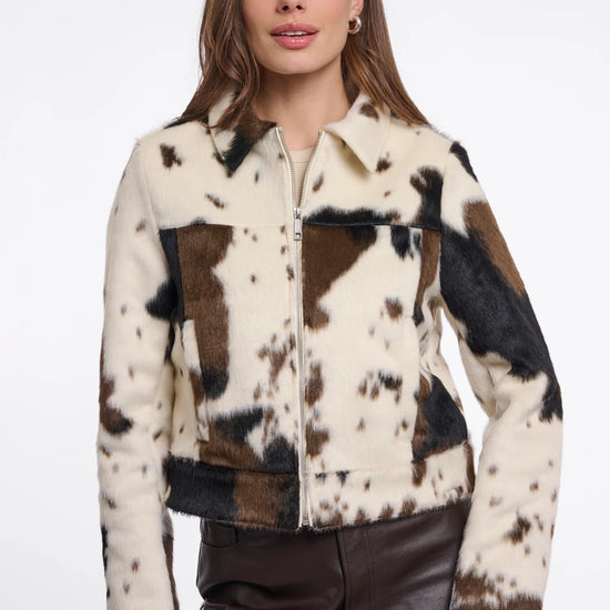 A woman modeling a cropped faux-fur jacket with a cow-print pattern in cream, brown, and black, paired with dark brown faux-leather pants against a plain white studio background.