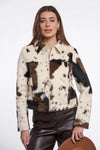 A woman modeling a cropped faux-fur jacket with a cow-print pattern in cream, brown, and black, paired with dark brown faux-leather pants against a plain white studio background.