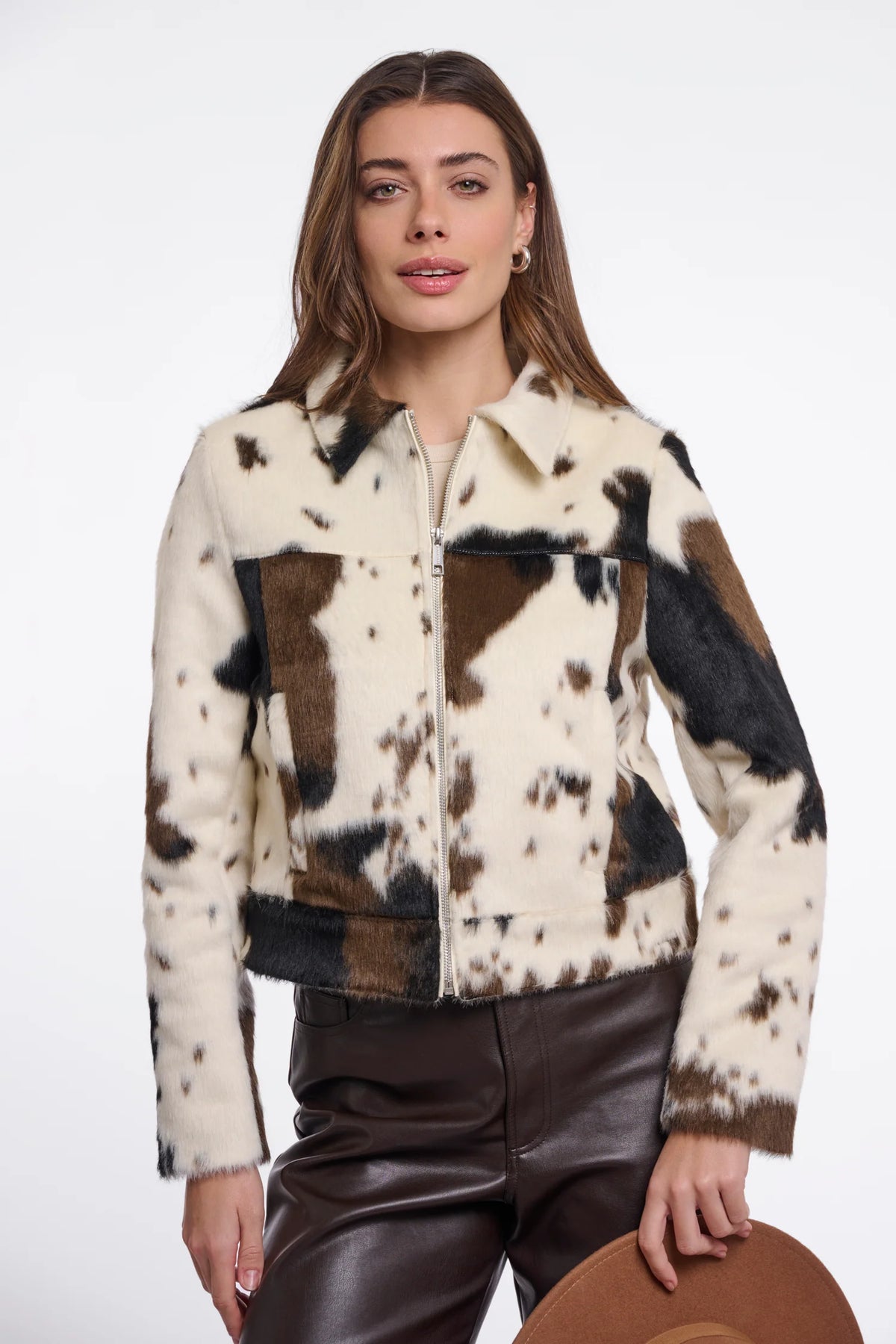 A woman modeling a cropped faux-fur jacket with a cow-print pattern in cream, brown, and black, paired with dark brown faux-leather pants against a plain white studio background.