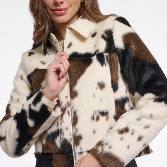 A woman wearing a cropped faux-fur jacket with a cow-print pattern in cream, brown, and black, paired with dark brown pants, against a plain white studio background.