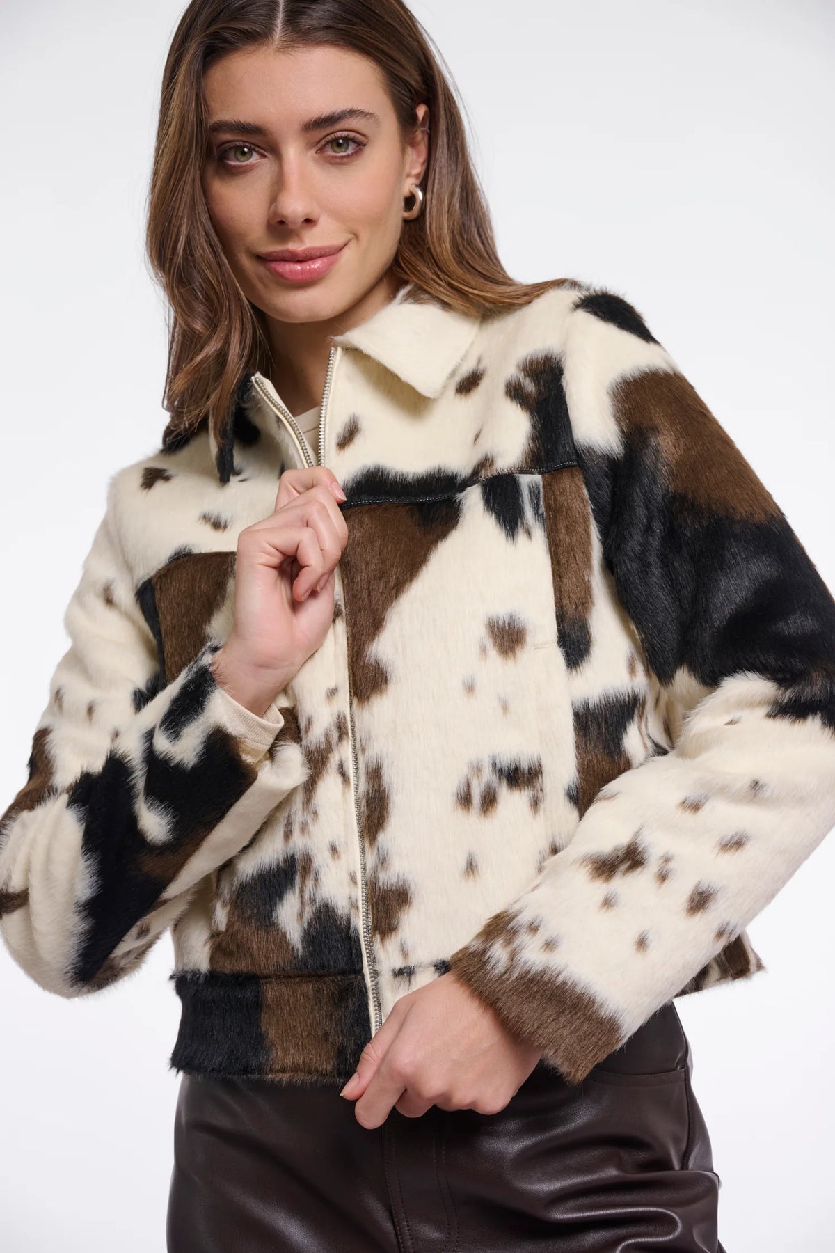 A woman wearing a cropped faux-fur jacket with a cow-print pattern in cream, brown, and black, paired with dark brown pants, against a plain white studio background.