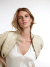 A woman with blonde hair wearing a beige structured jacket over a white satin top, accessorized with gold jewelry, photographed against a clean white background.